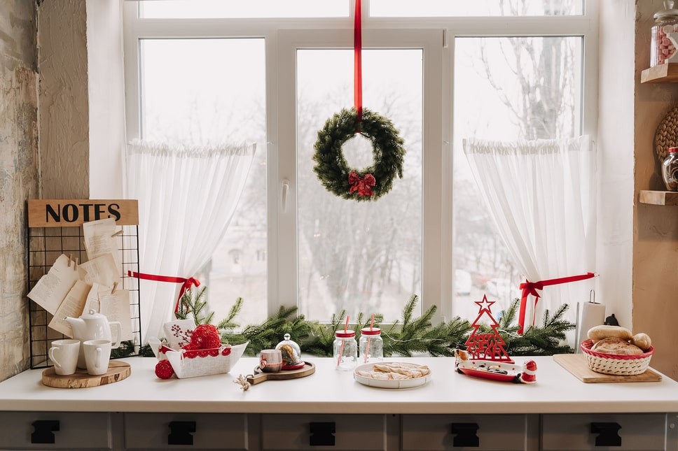 A kitchen counter bar area covered with Christmas treats and windows decorated with sheer curtains a