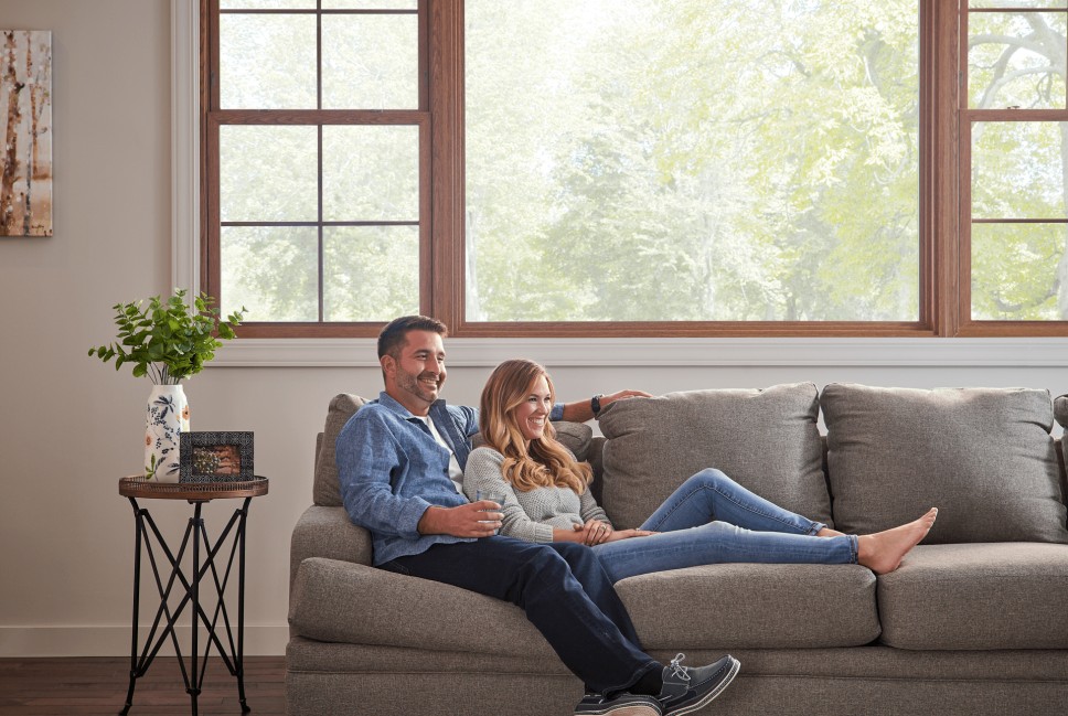 Man and woman smiling as they relax on a gray sofa in a naturally lit room with large picture window