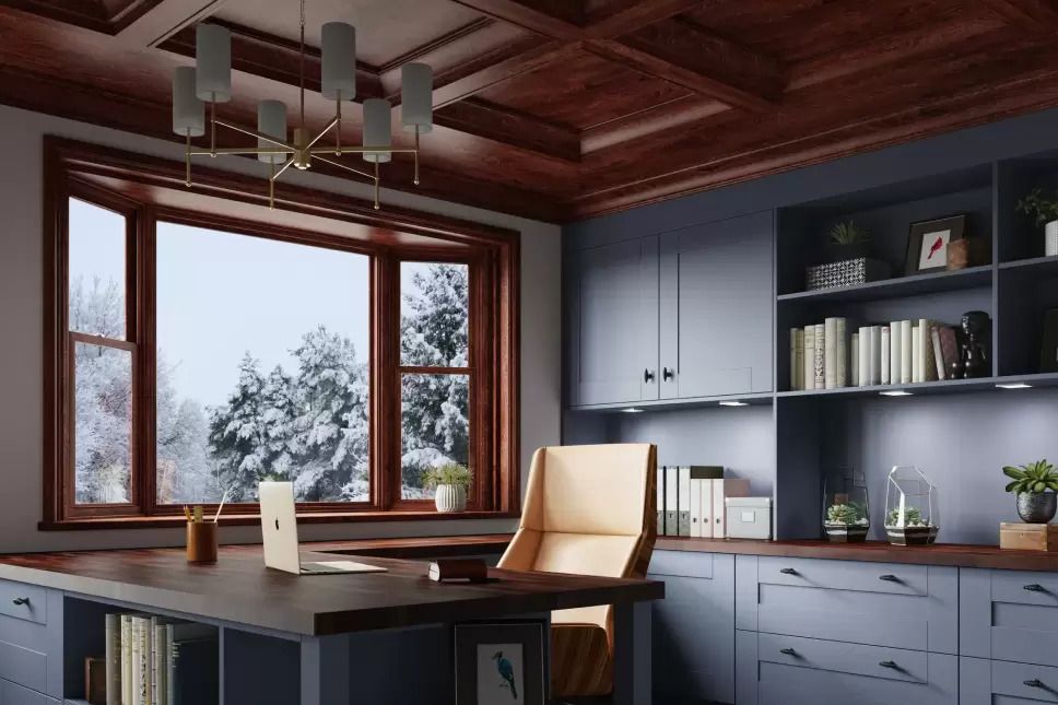 Home office with a wood-trimmed bay window overlooking snow-covered evergreens on a winter day.